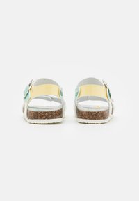 Bakvy av sandaler med en gul och vit design, justerbara remmar, korksula och turkosa accenter. Platt, avslappnad stil.