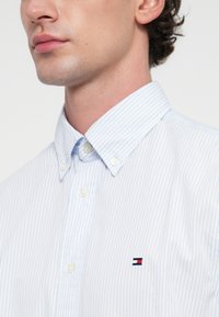 Λευκό και ανοιχτό μπλε ριγέ πουκάμισο με κολάρο, που διαθέτει ένα μικρό κεντημένο λογότυπο στο στήθος και στρογγυλά, λευκά κουμπιά.