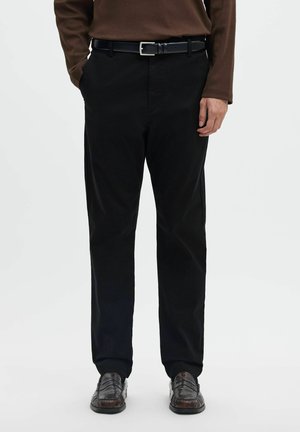 Homme portant un pantalon noir à jambes droites, une ceinture marron, un haut marron à manches longues et des chaussures en cuir marron foncé, debout devant un fond uni.