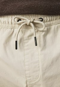 Beige rövidnadrág, húzózsinórral az derékrészen, texturált kötéllel és fekete műanyag végződéssel, sima szövetfelülettel.