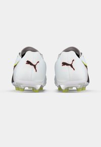 Białe korki piłkarskie z czarno-czerwonym logo, żółtymi akcentami i teksturowanym wzorem, z wyraźnymi, kolczastymi podeszwami zapewniającymi przyczepność.