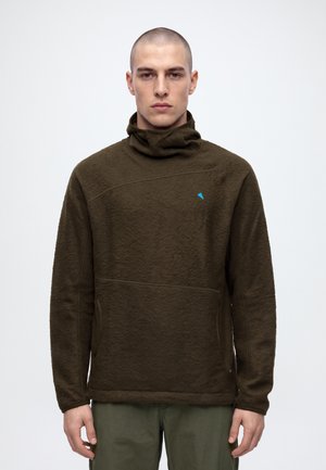 Un homme se tient debout face à l’objectif, vêtu d’un pull texturé vert olive foncé à col cheminée montant et d’un pantalon assorti, sur un fond clair uni.