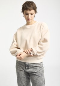 Jasno beżowy polarowy sweter z okrągłym dekoltem, ściągaczami przy mankietach i dole. Model nosi szare dżinsy z efektem acid-wash i ma krótkie włosy.