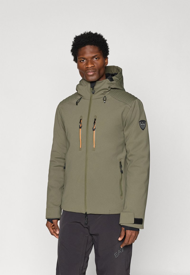 Olivovo zelená nepremokavá bunda s kapucňou, dvomi náprsnejami s oranžovými zipsami a rebrovanými detailami na pleciach. Prítomné čierne akcenty.