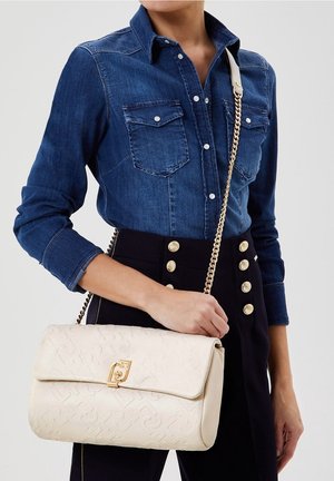 Kobieta ubrana w niebieską dżinsową koszulę i czarne spodnie z wysokim stanem oraz złotymi guzikami, trzymająca kremową teksturowaną torebkę na złotym łańcuszku.