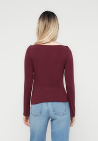 Bordeauxkleurige top met lange mouwen, een brede halslijn, een aansluitend ontwerp en een zachte textuur, gecombineerd met lichtblauwe denim jeans, gezien vanaf de achterkant.
