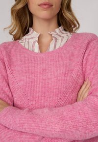 Pull en tricot rose avec une finition texturée et un col large et arrondi, superposé à une blouse blanche à rayures avec volants.