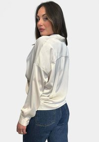 Blusa bianca in satin con maniche lunghe e vestibilità comoda, dotata di colletto con bottoni e dettaglio plissettato sul retro, abbinata a jeans blu scuro.