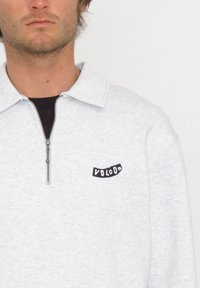 Suéter de forro polar gris claro con cremallera y cuello tipo chal, que presenta un logo bordado en negro en el pecho y un cierre de cremallera plateado.