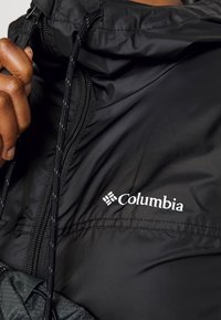 Černá bunda Columbia má vysoký límec, zapínání na zip a nastavitelné stahovací šňůrky. Výrazné logo je umístěno na oblasti hrudníku.