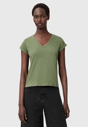 Camiseta verde de algodón con escote en V y mangas cortas enrolladas; corte relajado. Combinada con pantalones negros, diseño minimalista y textura suave.