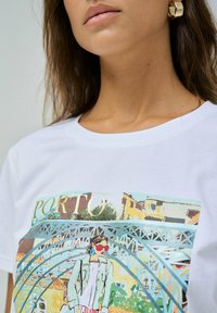 Camiseta de algodón blanca con un estampado gráfico colorido de una mujer frente a un puente, con la palabra "PORTO" en dorado sobre la imagen.