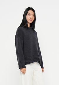 Junge Frau mit langen schwarzen Haaren, die einen oversize dunkelgrauen Pullover und weiße Hose trägt, steht vor einem schlichten weißen Hintergrund.