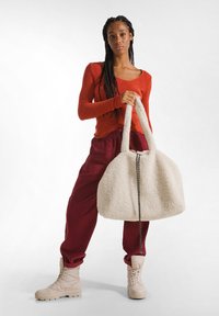 Witte pluche tas met twee handvatten, voorzien van een gestructureerd oppervlak en een zwart ritsdetail. Het model draagt een rode top en bordeauxrode broek.
