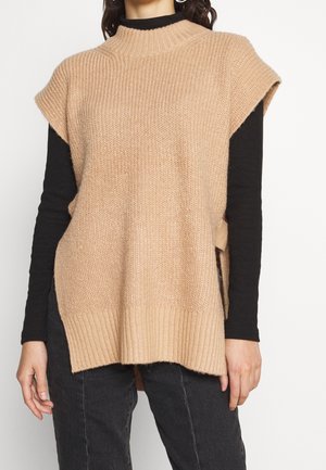 Mannequin portant un pull sans manches en maille beige par-dessus un haut noir à manches longues et un jean foncé.