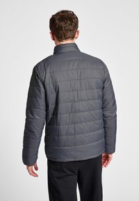 Man die een donkergrijs gewatteerd jack en zwarte broek draagt, met zijn rug naar voren staand tegen een eenvoudige lichte achtergrond.