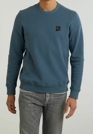 Homme portant un sweat-shirt bleu à col rond avec un logo patch noir sur la poitrine et un jean gris, debout devant un fond uni.