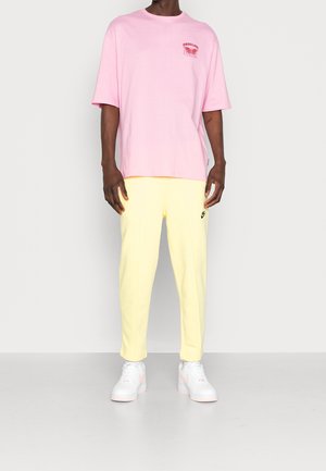 Roze katoenen T-shirt met korte mouwen en een rood grafisch motief op de borst, gecombineerd met gele joggingbroek en witte sneakers met roze accenten.