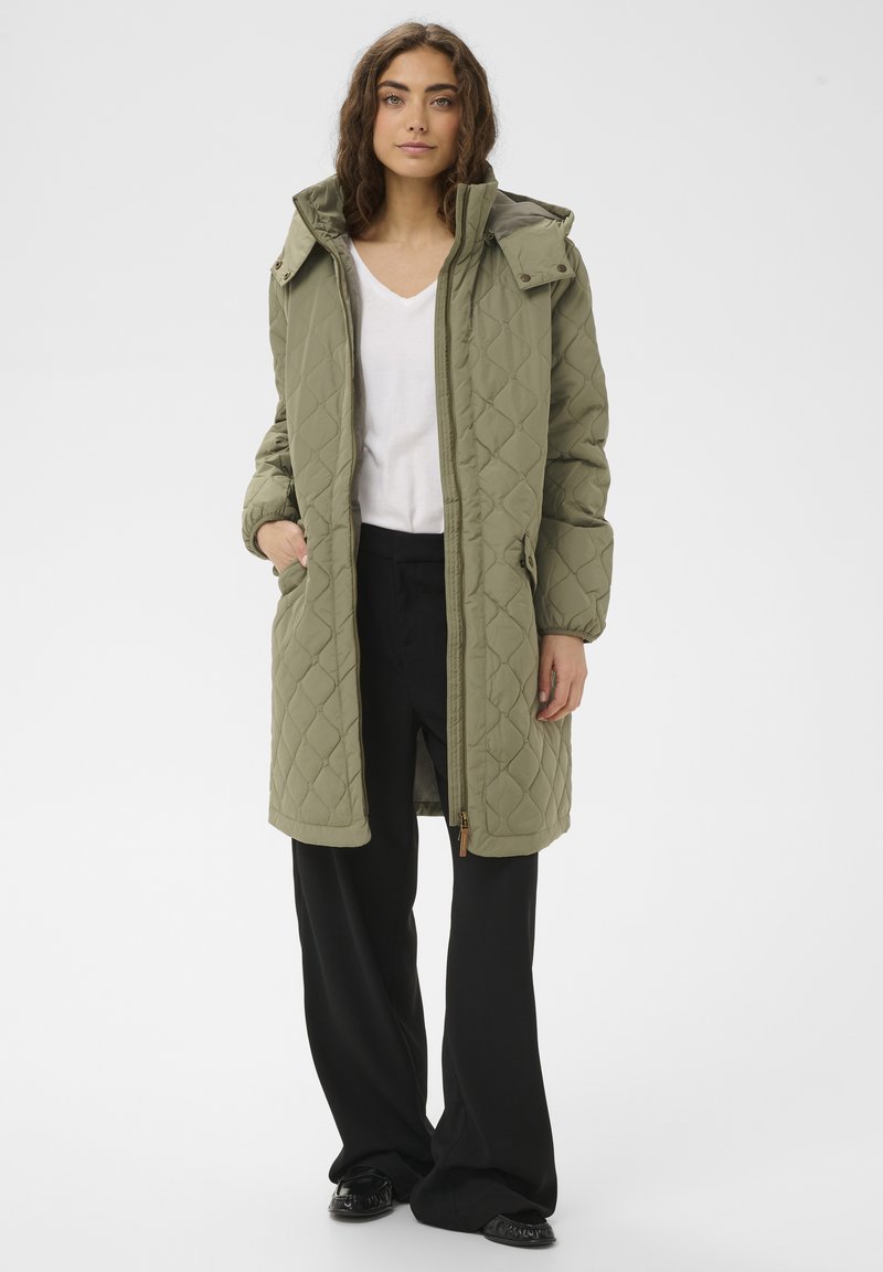 Chaqueta acolchada verde oliva con capucha, con cierre delantero, bolsillos laterales y puños acanalados. Se lleva sobre una camiseta blanca de cuello en V y pantalones negros.