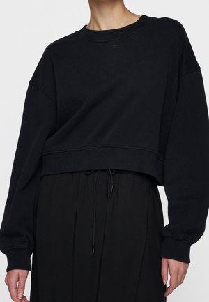 Person trägt ein schwarzes, kurz geschnittenes Sweatshirt mit langen Ärmeln und gerippten Bündchen, kombiniert mit schwarzen Jogginghosen mit Kordelzug.