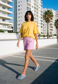 Jeune femme marchant dans une rue urbaine ensoleillée, portant un t-shirt jaune, un short rose et des sandales bleues, avec en arrière-plan des bâtiments blancs modernes et des palmiers.