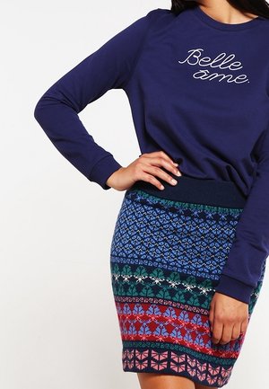 Kobieta ubrana w granatową bluzę z napisem "Belle âme" oraz kolorową, wzorzystą dzianinową spódnicę, z ręką opartą na talii.