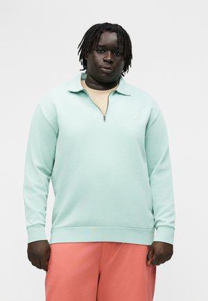 Άτομο με σκουρόχρωμο δέρμα και κοντά dreadlocks φοράει ανοιχτό μπλε πουλόβερ με φερμουάρ και κοραλλί παντελόνι σε απλό φόντο.