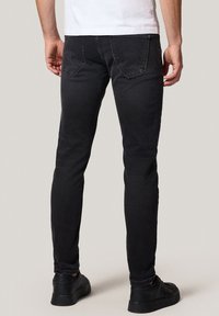 Mężczyzna w czarnych, dopasowanych dżinsach i czarnych sneakersach, stojący z rękami swobodnie opuszczonymi wzdłuż ciała na jednolitym tle.