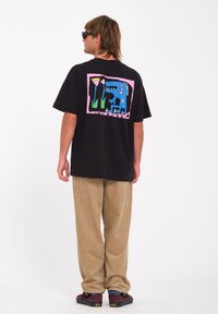 Zwarte katoenen t-shirt met een kleurrijk grafisch ontwerp op de achterkant met een blauwe dinosaur, bloemen en een roze kader. Gecombineerd met beige corduroy broek.