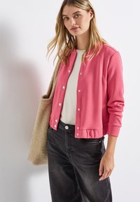 Růžová mikina s knoflíky, žebrovaným lemem a manžetami, v kombinaci s krémovou košilí a tmavě šedými kalhotami. Modelka drží béžovou tašku.
