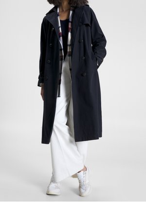 Person iført en mørkeblå trenchcoat, hvide vidde bukser, hvide sneakers og en ternet skjorte synlig under frakken.