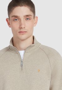 Beige rits sweatshirt met een hoge kraag, voorzien van een zilveren rits en een klein oranje logo. Glad oppervlak en raglanmouwen.