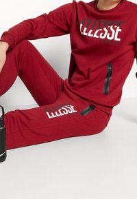 Czerwona sportowa odzież składająca się z bluzy i spodni, obie z zamykanymi kieszeniami. Na górze widnieje logo "Ellesse" w wyraźnym napisie.