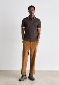 Braunes Polohemd mit weißem Besatz, kombiniert mit orangen Cordhosen. Die Schuhe sind grüne Turnschuhe. Einfaches, lässiges Passform-Design.