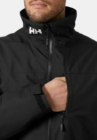 Chaqueta negra con cuello alto, cierre frontal y puños de velcro. Cuenta con un bolsillo en el pecho y tejido texturizado. Logotipo mostrado en el cuello.