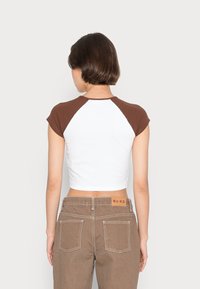 Kvinna med kort brunt hår står med ryggen mot oss, iförd en vit crop top med bruna ärmar och högmidjade bruna byxor.