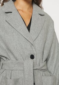 Vrouw in een grijze wollen, met riem afgewerkte jas met brede revers, met één zwart knoopje vastgemaakt, en zwarte kleding zichtbaar bij de halslijn.