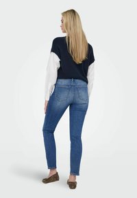 Un pull en maille marine superposé sur une chemise blanche, associé à un jean bleu ajusté. Le jean présente des poches arrière et un léger délavage. Des mocassins marron aux pieds.