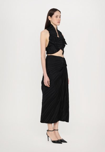 Black pleated two-piece outfit featuring a cropped top with ruffle accents and a long, draped skirt, paired with black stiletto heels.