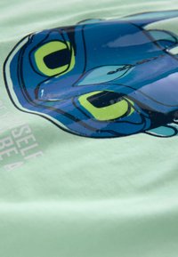 Mintgrön t-shirt med ett glansigt blått och gult ansikte av en tecknad karaktär, med tryckt text nedanför i en ljus font.