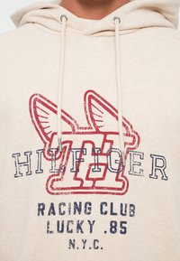 Zbliżenie beżowej bluzki z kapturem z czerwonym logo w postaci skrzydlatej litery "H" oraz ciemnoniebieskim napisem "Hilfiger Racing Club Lucky 85 N.Y.C."