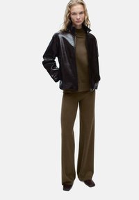 Črna sijoča usnjena jakna z visokim ovratnikom, v kombinaciji s temno olivno zelenim pletenim puloverjem in usklajenimi širokimi hlačami. Rjave čevlje.
