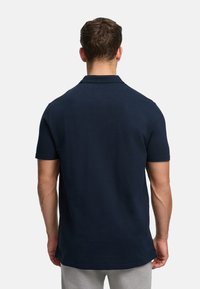 Mies, joka käyttää yksiväristä tummansinistä lyhythihaista poolopaitaa, nähtynä takaa vaaleaa taustaa vasten.