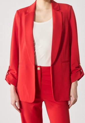 Femme portant un blazer rouge vif à manches retroussées, un pantalon rouge assorti et un haut blanc à col carré sous le blazer.