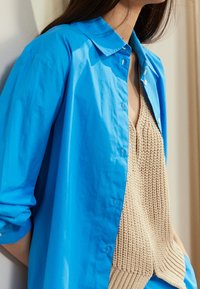 Blue button-up shirt made of lightweight material, layered over a beige knitted sweater. The shirt has a classic collar and rolled sleeves.
