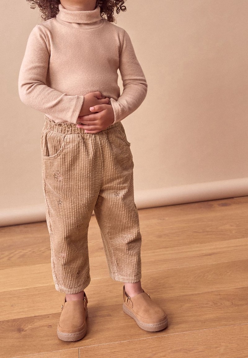 Enfant aux cheveux bouclés portant un col roulé beige, un pantalon en velours côtelé et des chaussures sans lacets, debout sur un sol en bois, les mains posées sur le ventre.
