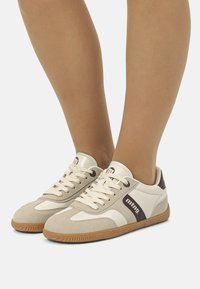 Zapatillas de deporte crema con acentos de ante gris, lengüeta acolchada y cordones redondos. Presentan una suela de goma marrón y un detalle del logo oscuro en el costado.