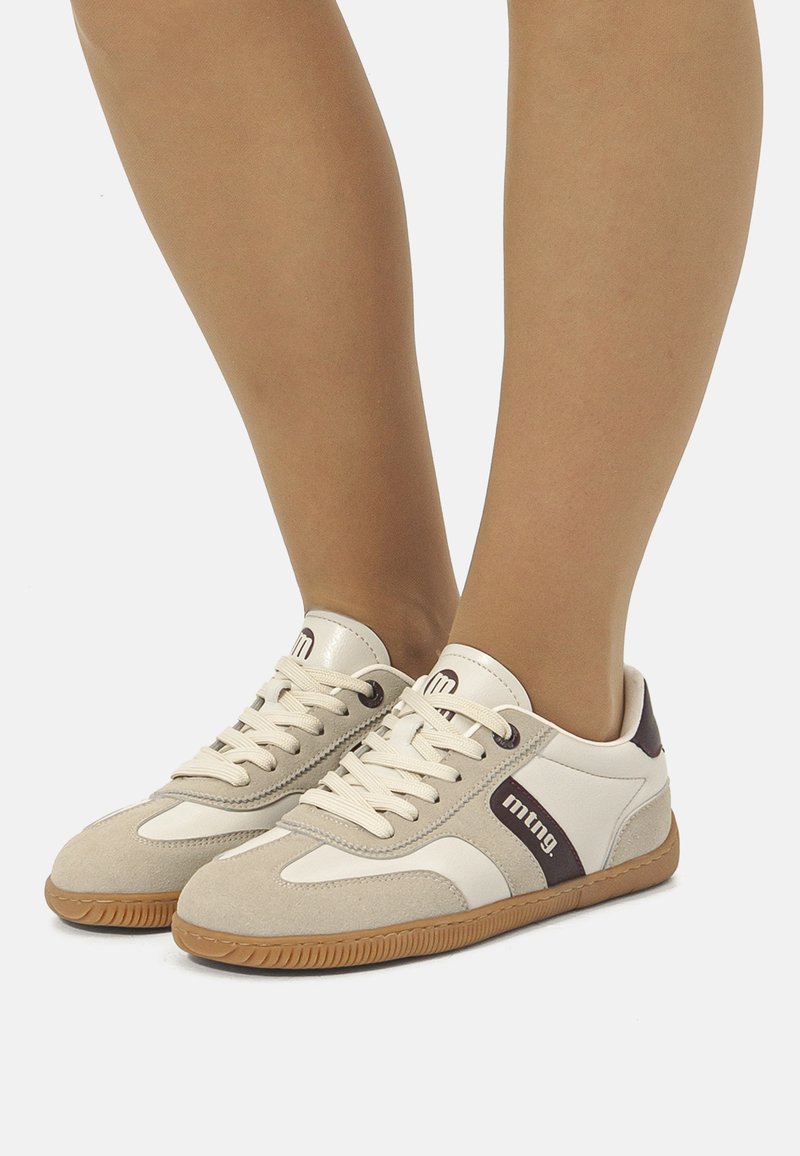 Zapatillas de deporte crema con acentos de ante gris, lengüeta acolchada y cordones redondos. Presentan una suela de goma marrón y un detalle del logo oscuro en el costado.