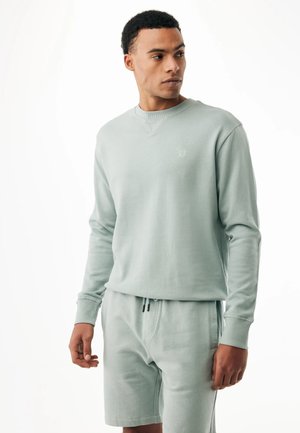 Uomo che indossa una felpa a maniche lunghe verde chiaro e pantaloncini abbinati, che guarda verso sinistra su uno sfondo bianco semplice.