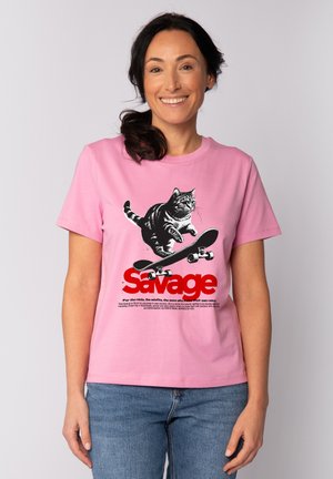 Vrouw die glimlacht en een roze T-shirt draagt met een zwart-witte kat die op een skateboard springt en rode tekst "Savage" aan de voorkant.